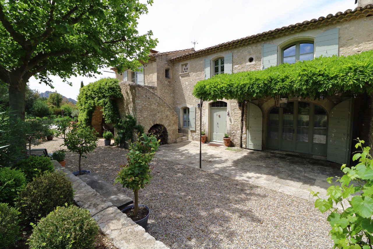 A picturesque view of a traditional french farmhouse with stone walls, green shutters, and a landscaped garden with neatly trimmed trees and gravel pathways.